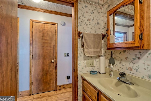 bathroom with a textured ceiling, wood finished floors, wallpapered walls, baseboards, and vanity