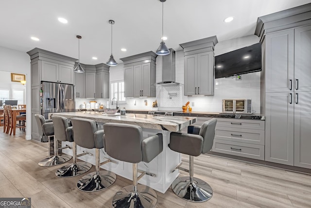 kitchen featuring light stone counters, gray cabinetry, decorative backsplash, wall chimney range hood, and stainless steel fridge