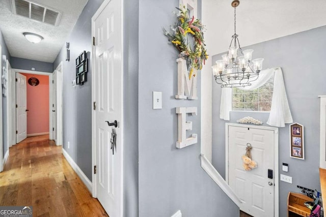 hall featuring visible vents, baseboards, wood finished floors, a notable chandelier, and a textured ceiling