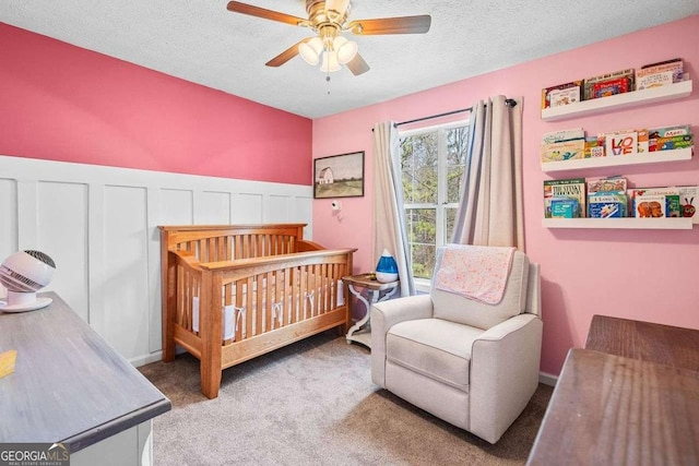 carpeted bedroom featuring a ceiling fan, a nursery area, wainscoting, and a textured ceiling