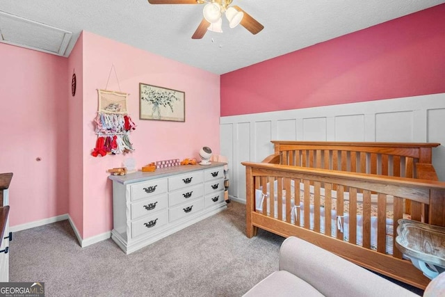 carpeted bedroom with a crib, a ceiling fan, a textured ceiling, wainscoting, and attic access