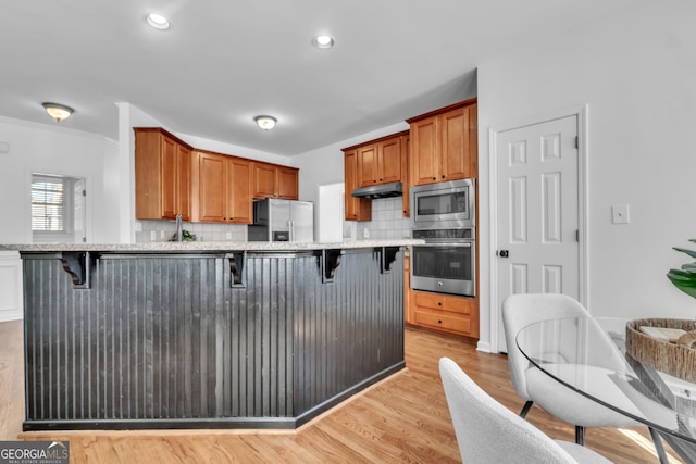 1654 Summersweet Ln, Dacula, GA 30019 - kitchen with appliances with stainless steel finishes, under cabinet range hood, a kitchen breakfast bar, decorative backsplash, and light wood-type flooring