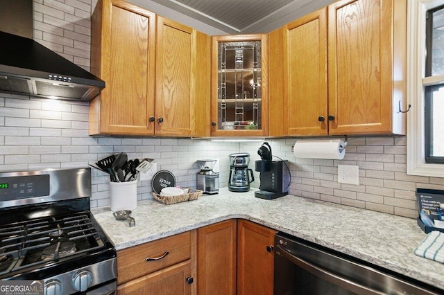 kitchen featuring backsplash, glass insert cabinets, light stone counters, appliances with stainless steel finishes, and wall chimney exhaust hood