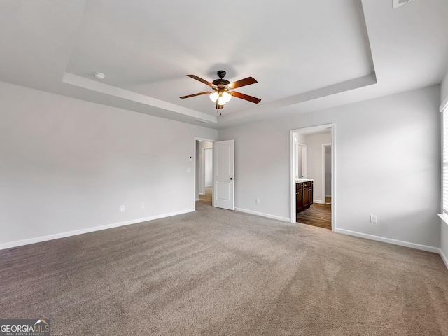 unfurnished bedroom with a ceiling fan, a tray ceiling, dark carpet, connected bathroom, and baseboards