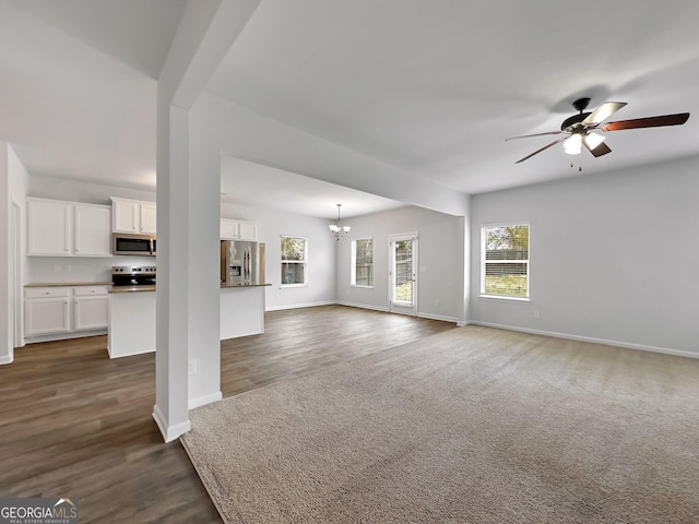 unfurnished living room featuring dark carpet, ceiling fan with notable chandelier, dark wood-type flooring, and baseboards