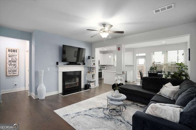 living area with visible vents, a glass covered fireplace, dark wood-style floors, baseboards, and ceiling fan