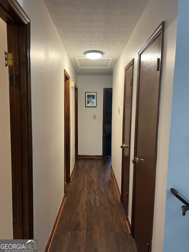 hall with a textured ceiling, attic access, dark wood-type flooring, and baseboards