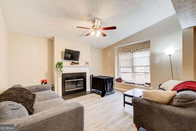 living room with a textured ceiling, a glass covered fireplace, light wood finished floors, lofted ceiling, and ceiling fan