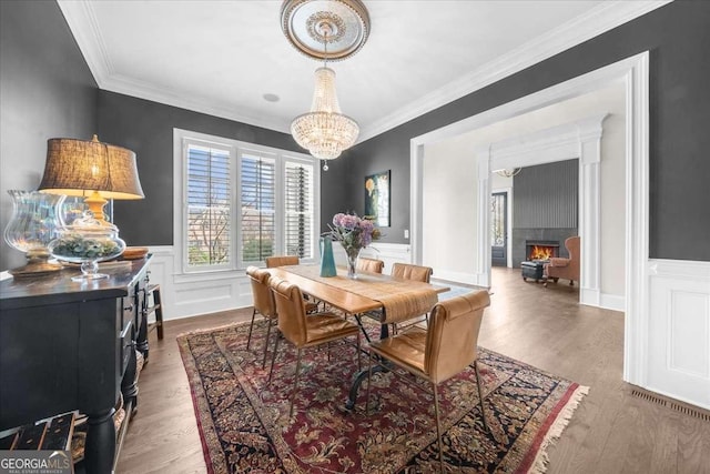 dining area with a wainscoted wall, ornamental molding, wood finished floors, a lit fireplace, and a chandelier