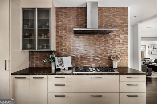 kitchen featuring dark stone counters, stainless steel gas stovetop, glass insert cabinets, wall chimney exhaust hood, and tasteful backsplash