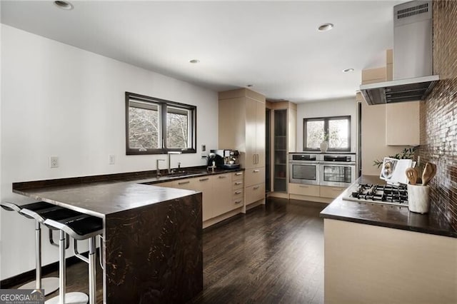 kitchen with a peninsula, a sink, appliances with stainless steel finishes, dark countertops, and wall chimney range hood