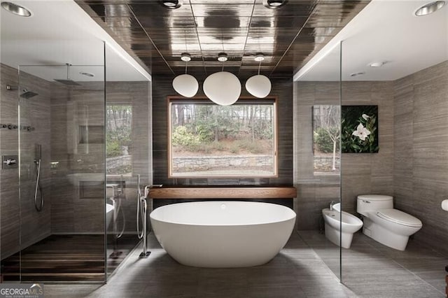 bathroom featuring a freestanding tub, toilet, a stall shower, a bidet, and tile walls