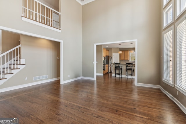 unfurnished living room with visible vents, baseboards, stairs, a towering ceiling, and dark wood-style flooring