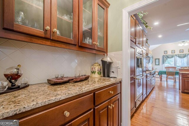 kitchen with light stone counters, light wood-type flooring, decorative backsplash, glass insert cabinets, and brown cabinets