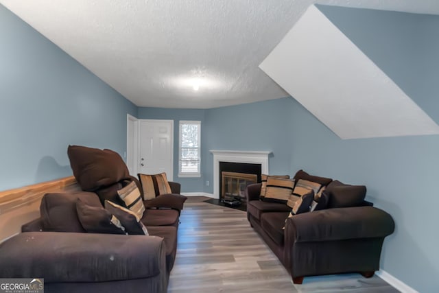 living room featuring baseboards, a fireplace with flush hearth, lofted ceiling, wood finished floors, and a textured ceiling