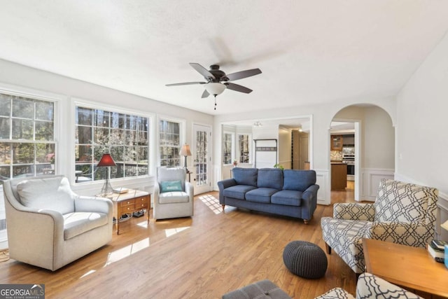 living room featuring a wainscoted wall, light wood-style flooring, arched walkways, ceiling fan, and a decorative wall