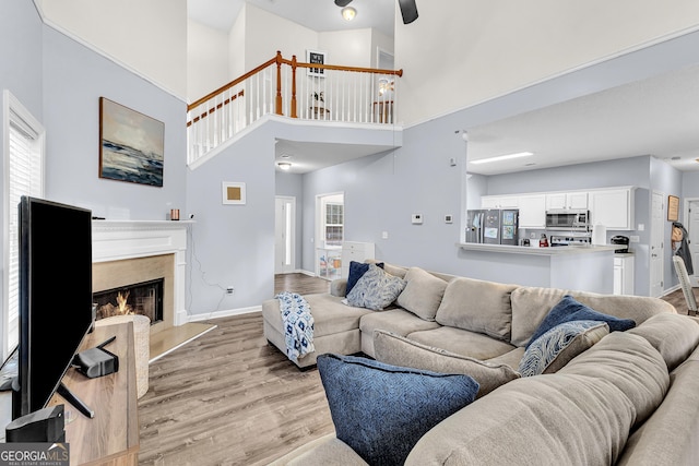 living area featuring baseboards, a fireplace, ceiling fan, light wood-style floors, and a towering ceiling