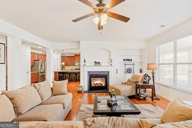 living room with built in features, a ceiling fan, baseboards, a glass covered fireplace, and light wood-type flooring