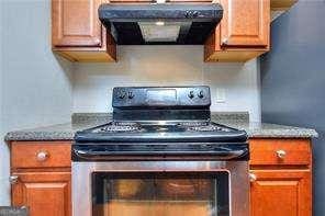 kitchen with range hood, stone counters, stainless steel electric stove, freestanding refrigerator, and brown cabinets