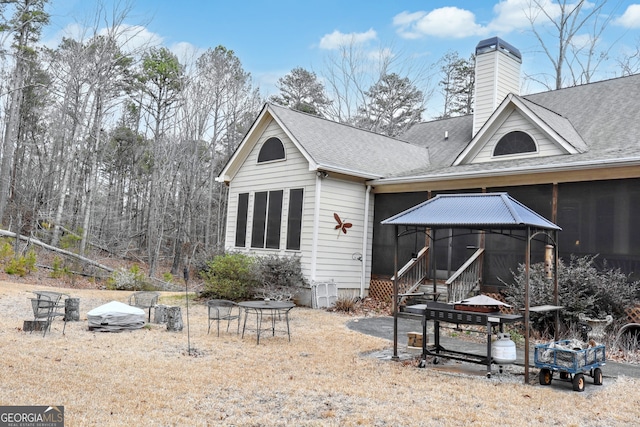 back of house with a patio, roof with shingles, a sunroom, and a chimney