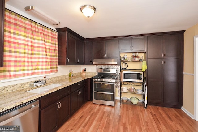 kitchen with light wood-style flooring, under cabinet range hood, a sink, stainless steel appliances, and dark brown cabinetry