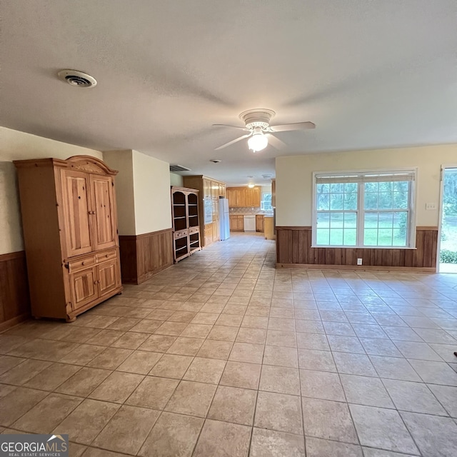 unfurnished living room with visible vents, wooden walls, wainscoting, light tile patterned floors, and ceiling fan