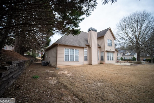 rear view of house featuring a shingled roof, central AC, a lawn, a chimney, and a patio