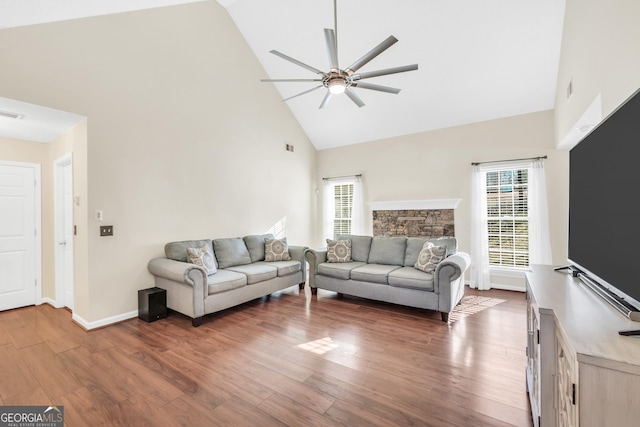 living room with a ceiling fan, wood finished floors, baseboards, high vaulted ceiling, and a stone fireplace