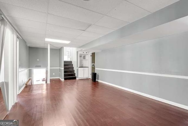 finished basement featuring a paneled ceiling, baseboards, dark wood-style flooring, and stairs