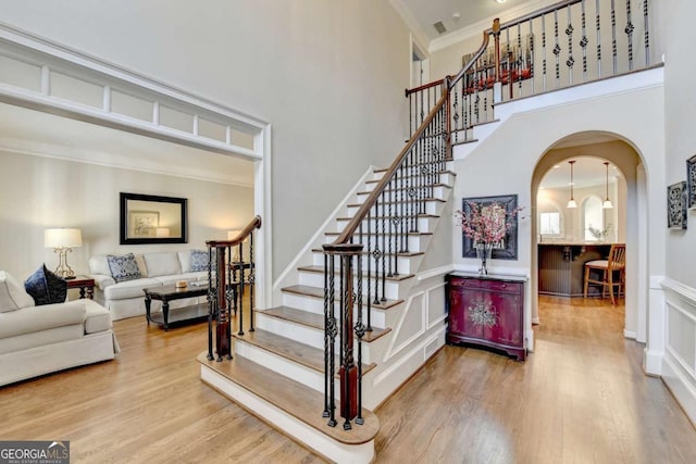 staircase with ornamental molding, wood finished floors, arched walkways, wainscoting, and a towering ceiling