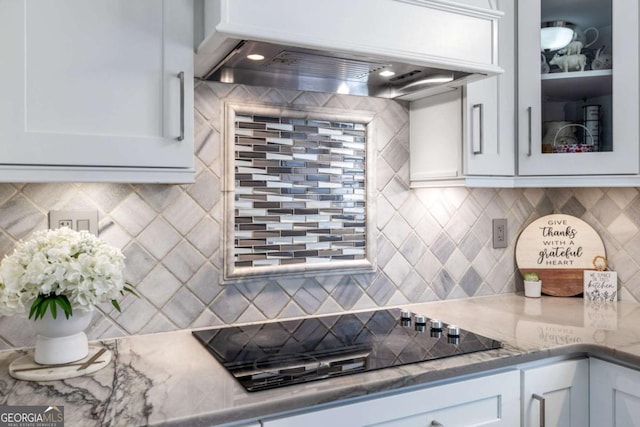 kitchen featuring decorative backsplash, custom range hood, glass insert cabinets, white cabinets, and black electric cooktop