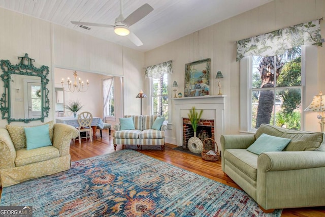 living area with ceiling fan with notable chandelier, a brick fireplace, wood finished floors, and visible vents