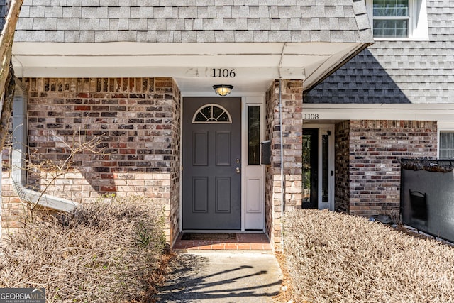 doorway to property featuring brick siding and roof with shingles