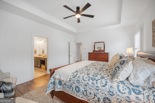 bedroom with a raised ceiling, light wood-style flooring, ensuite bathroom, a ceiling fan, and baseboards