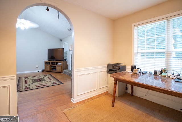 home office with vaulted ceiling, light wood-style flooring, wainscoting, arched walkways, and a textured ceiling