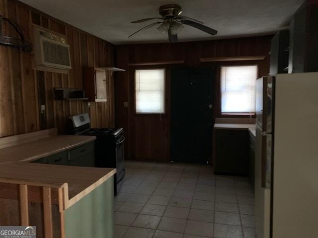 kitchen with freestanding refrigerator, wood walls, light tile patterned flooring, gas range, and ceiling fan