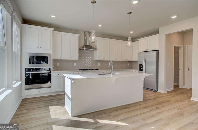 kitchen with a sink, light countertops, appliances with stainless steel finishes, wall chimney range hood, and light wood-type flooring