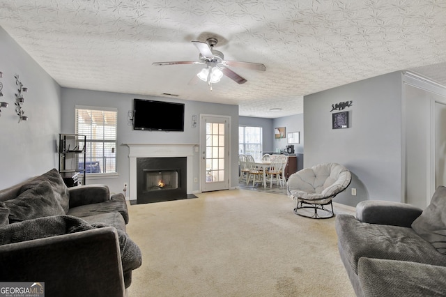 carpeted living area featuring a wealth of natural light, a fireplace with flush hearth, a textured ceiling, and a ceiling fan