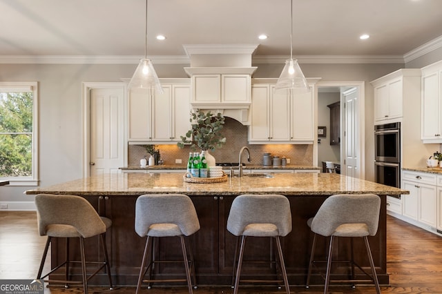 kitchen featuring a kitchen island with sink, a sink, white cabinetry, stainless steel double oven, and dark wood-style flooring