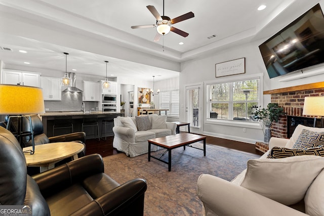 living room featuring visible vents, a brick fireplace, ceiling fan with notable chandelier, a raised ceiling, and dark wood-style flooring