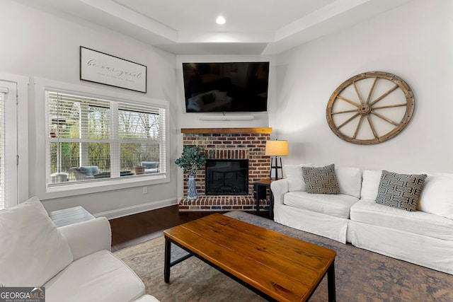 living area featuring a brick fireplace, baseboards, a tray ceiling, recessed lighting, and wood finished floors