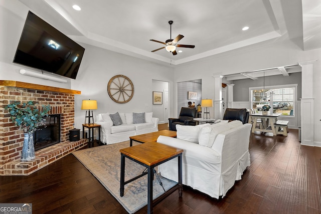 living room with decorative columns, ceiling fan, dark wood-type flooring, a raised ceiling, and a brick fireplace