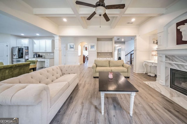 living area with beam ceiling, light wood-style flooring, arched walkways, coffered ceiling, and a ceiling fan