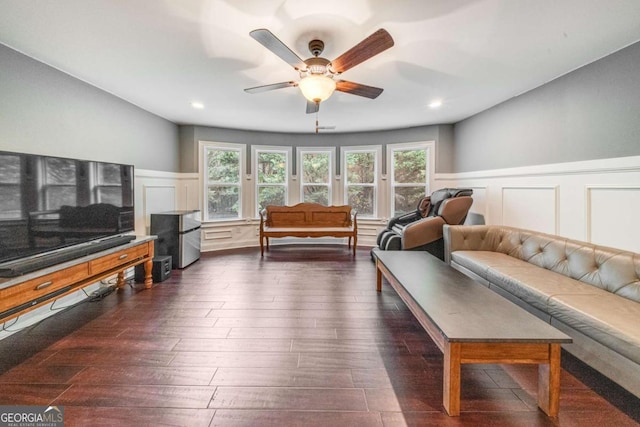 living room featuring dark wood finished floors, a decorative wall, a ceiling fan, and a wainscoted wall