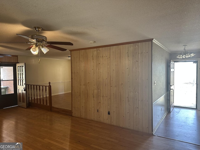 empty room featuring wood walls, ornamental molding, ceiling fan with notable chandelier, wood finished floors, and a textured ceiling