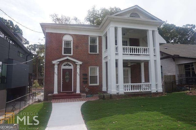 greek revival inspired property with a front yard, a balcony, fence, covered porch, and brick siding