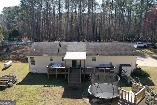 rear view of house with a wooden deck, a trampoline, roof with shingles, and a lawn