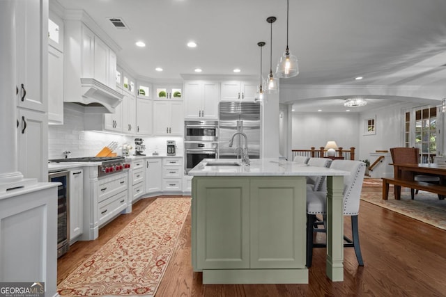 kitchen featuring beverage cooler, a kitchen island with sink, a sink, stainless steel appliances, and white cabinets