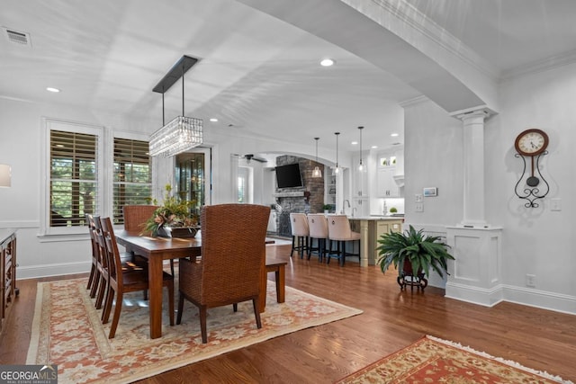 dining space with visible vents, crown molding, a stone fireplace, wood finished floors, and ornate columns