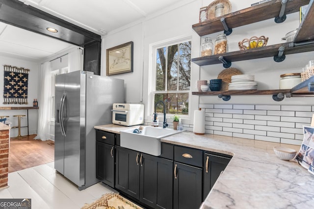 kitchen with dark cabinetry, open shelves, a sink, stainless steel fridge, and backsplash